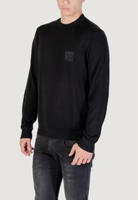 Maglione nero a maglia con collo rotondo, polsini e orlo a coste, che presenta una patch testurizzata nera con logo sul petto.
