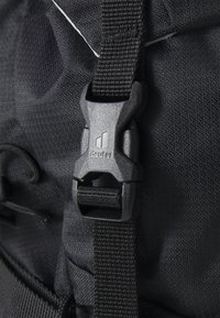 Rucsac din material textil negru, în prim-plan, cu un cataramă din plastic ce poartă logo-ul "Deuter", cusături întărite și o suprafață texturată.