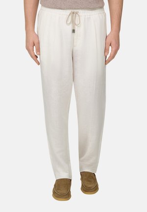 Hvide herre-linnedbukser med snøre i taljen, kombineret med brune ruskinds-mokassinsko og en beige top, stående mod en ensfarvet baggrund.