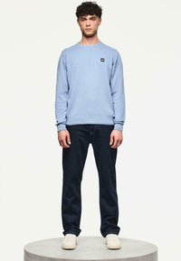 Uomo in piedi su una piattaforma rotonda, indossa un felpa azzurro chiaro, jeans scuri e sneaker bianche, con un'espressione neutra su uno sfondo chiaro.