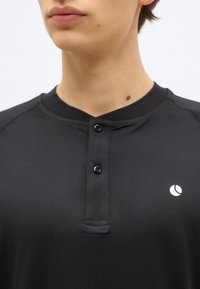 Κοντινή λήψη ενός ατόμου που φοράει μαύρο πόλο με κουμπιά και ένα μικρό λευκό κυκλικό λογότυπο στο στήθος.