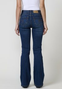 Jeans acampanados de un azul oscuro, hechos de denim, que presentan una cintura alta, costuras intrincadas en los bolsillos traseros y una etiqueta de cuero en la cintura.