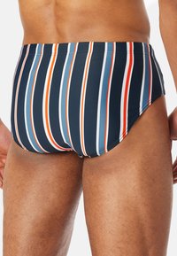 Simlabbare i en marinblå bakgrund med vertikala ränder i orange, vitt och blått. Tillverkade av ett mjukt, stretchigt material.