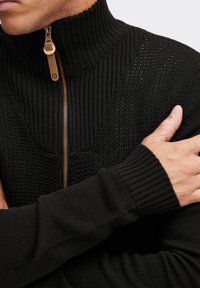 Schwarzer Reißverschluss-Pullover mit hohem Rippkragen, strukturiertem Strickmuster und kontrastierendem bronzefarbenem Reißverschluss mit Ledergriff.