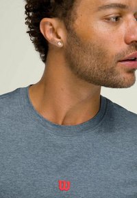 Camiseta de color gris heather con cuello redondo y un pequeño logo rojo de "W" en el pecho. La tela parece suave y ligeramente texturizada.