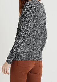 Frau in einem schwarz-weiß melierten Strickpullover und rostfarbigen Hosen, seitlich und von hinten vor einem einfarbigen Hintergrund gezeigt.