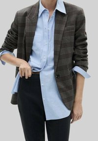 Hnědý károvaný blazer přes světle modrou košili s knoflíky, s uvolněným střihem a dobře padnoucím designem. Tmavé kalhoty doplňují outfit.