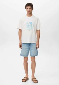 Hvid bomulds t-shirt med et blåt grafisktryk med fugle, parret med lyse denimshorts og brune sandaler. Enkel, afslappet design.