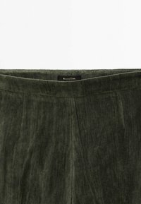 Pantalon en velours vert foncé avec une taille haute et des plis discrets à l'avant, étiqueté avec une étiquette noire à l'intérieur de la taille.