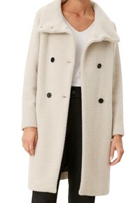 Femme portant un manteau en laine beige à chevrons longueur genoux avec des boutons noirs, associé à un haut blanc et un pantalon noir.