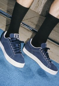 Marina mockasiner av mocka med vita gummisulor, rund tå och svarta snören. Svarta ribbade strumpor delvis synliga, mot en blå bakgrund.