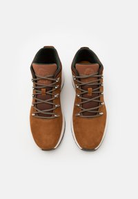 Zapatos de senderismo de ante marrón con detalles en verde oscuro, que presentan herrajes de metal, cordones grises y un detalle de lengüeta de montañismo con el logo grabado.