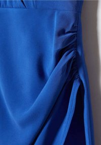 Niebieska satynowa sukienka z marszczeniami z boku. Gładka tekstura, dopasowany krój i ukośne cięcie, które podkreśla lekkość i opadanie materiału.