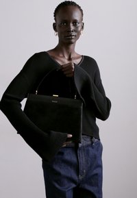 Melna ādas rokassoma ar strukturētu formu un zelta tonī piekabi. Modelis valkā melnu ribotu džemperi un zilos džinsus.