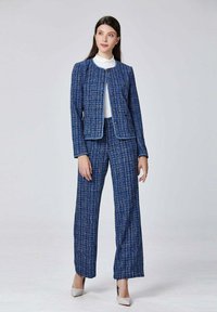 Costume en tweed bleu avec une veste ajustée, col rond et boutons dorés, associé à un pantalon à jambe large. Texture lisse, détails de motif subtils.