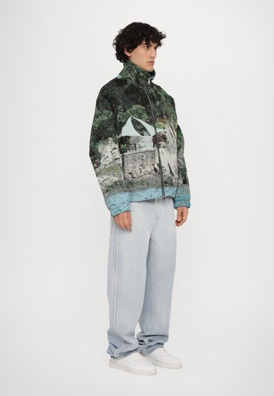 Zippered jacket featuring a landscape print with greenery and a building; made of fabric with soft texture. Paired with light blue jeans and white shoes.