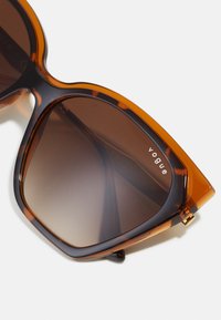 Lunettes de soleil dégradées brunes avec un design surdimensionné en œil de chat. Les montures présentent un motif écaille de tortue avec des accents dorés. Le texte indique "vogue."