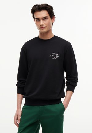 Νέος άνδρας που φοράει ένα μαύρο φούτερ Tommy Hilfiger και πράσινο παντελόνι, στέκεται με το ένα χέρι στην τσέπη μπροστά σε λευκό φόντο.