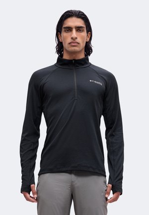 Mann mit schwarzem Columbia Langarm-Zip-Top und hellgrauer Hose steht vor einfachem hellem Hintergrund.