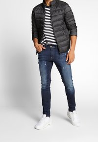 Schwarze Steppjacke mit gesteppter Textur, gestreiftes T-Shirt und dunkelblaue, verwaschene Skinny Jeans. Weiße Sneaker rundet das Outfit ab.