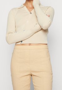 Personne portant un cardigan court crème à boutons mélangés et un pantalon beige taille haute, les bras croisés, montrant le ventre et une chaîne au cou.