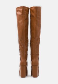 Bottes en cuir marron hautes avec un design élégant, dotées d'un bout arrondi et d'un talon épais, mises en avant par des fermetures éclair sur le côté. Texture lisse sur toute la surface.