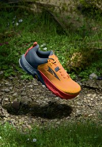 Chaussure de trail de course orange et grise avec un dessus texturé, un col rembourré, une semelle colorée et des accents réfléchissants, suspendue au-dessus de l'herbe.