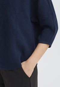 Pull en maille bleu marine avec un motif texturé, coupe ample, manches trois-quarts, associé à un pantalon noir et une main dans la poche.