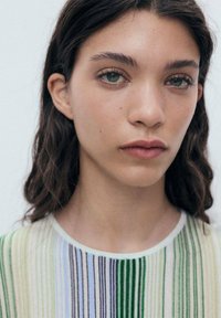 Primo piano di una persona con capelli scuri e mossi, che indossa un indumento a strisce multicolori con tonalità di verde, blu e bianco su uno sfondo chiaro.
