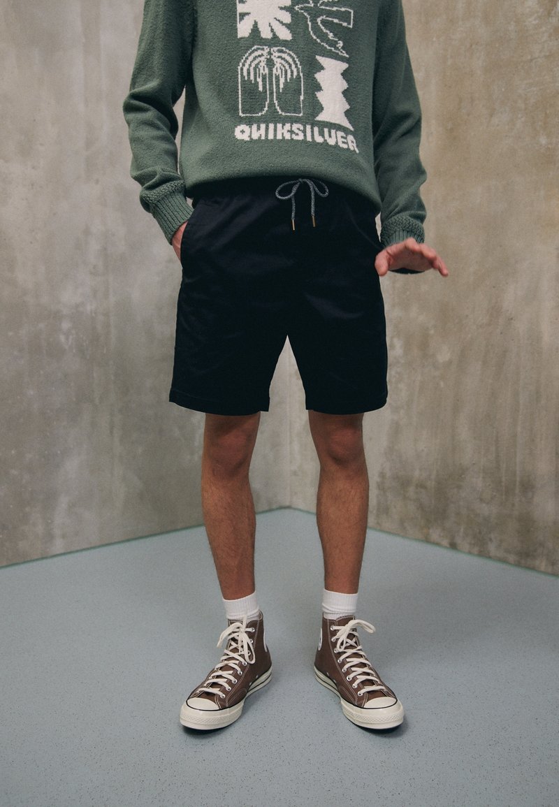 Personne portant un pull vert avec un graphisme blanc, un short noir, des chaussettes blanches et des baskets montantes marron, debout sur un sol gris contre un mur en béton.