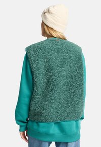 Vihreä fleece-liivi, jossa on teksturoitu pinta, kerroksittain teallin värisen collegepuseron päällä. Pipo on luonnonvalkoinen, jossa on resoripintakuvio. Rentoa asua.