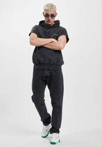 Svart hettegenser med korte ermer, kombinert med mørk grå jeans og hvite joggesko med grønne detaljer. Modell står med armene i kors.