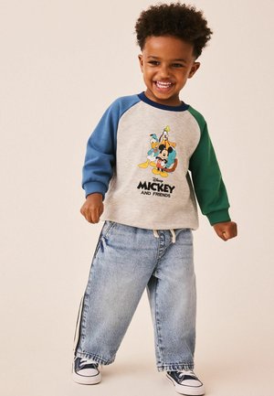 Smilende barn iført en grå sweatshirt med blå og grønne ærmer med Mickey og venner, løse jeans og sorte sneakers.