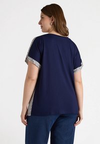 Marineblaues T-Shirt mit kurzen Ärmeln, das cremefarbene und marineblaue gemusterte Akzente am Saum aufweist. Weicher Stoff und entspannter Schnitt.