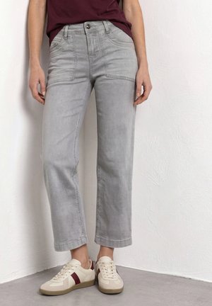 Personne portant un pantalon large court gris clair, une chemise bordeaux foncé et des baskets crème avec des rayures bordeaux, debout contre un mur blanc.
