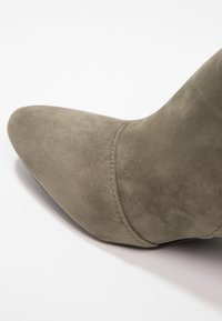 Bota de punta afilada en suave ante verde oliva, con una textura lisa y un sutil detalle de costura a lo largo de la costura.