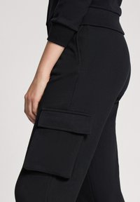 Czarne spodnie cargo z boczną kieszenią, prezentowane na dolnej części ciała osoby. Materiał wydaje się miękki i matowy. Brak widocznych wzorów.
