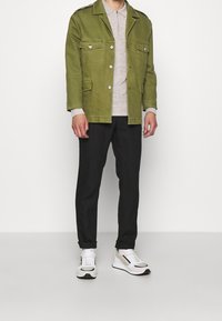 Zelená plátěná bunda s dvěma předními kapsami, šedé polo tričko, černé kalhoty a bílé a šedé tenisky, stojící na bílém pozadí.