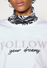 Camiseta blanca con texto embellecido con piedras brillantes que dice "FOLLOW" y texto en cursiva negra "your dreams", acompañada de una bufanda de cuello de patrón en blanco y negro.