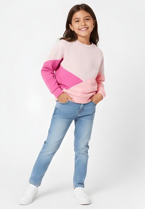 Uśmiechnięta młoda dziewczyna stoi z rękami w kieszeniach, ubrana w różową bluzę z blokami kolorów, jasnoniebieskie dżinsy i białe trampki na białym tle.