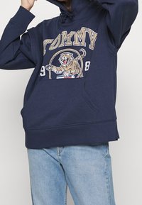 Námořnická modrá mikina s kapucí a šňůrkou, se grafickým motivem tygra a textem "TOMMY" ve zlaté barvě. Obsahuje před pockets a žebrované manžety.