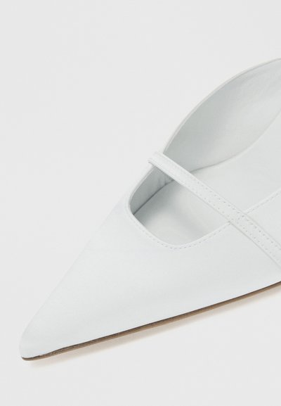 Sapato plano branco de bico fino com uma única alça fina na parte superior e sola castanho clara, sobre um fundo branco.