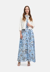 Witte denim kort jasje, gecombineerd met een lange blauwe jurk met een bloemenpatroon in tinten blauw en grijs, en witte blokhak sandalen.