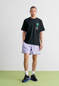 Svart grafisk t-skjorte med grønn kaktusprint, sammen med lyse lilla shorts. Svart sneakers med blå detaljer og hvite sokker.