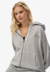 Joven mujer con cabello rubio vestida con una chaqueta con cremallera gris claro que dice "Superdry Athletics", posando contra un fondo blanco.
