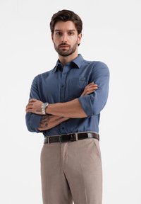 Camicia blu a maniche lunghe con bottoni per uomo, dotata di una tasca sul petto, abbinata a pantaloni beige chiaro e una cintura nera. Orologio argento al polso.