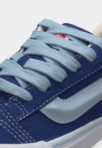 Μπλε αθλητικό παπούτσι με ανοιχτογάλαζα κορδόνια, λευκή λωρίδα στο πλάι και ορατό κόκκινο λογότυπο στη γλώσσα, εμφανίζεται σε κοντινή λήψη της μπροστινής άνω περιοχής.