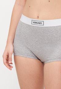 Szürke pamut boxer alsónadrág, fehér elasztikus derékkal, amelyen a "VERSACE" logó található. Közelkép a derékról és a nadrág oldaláról.