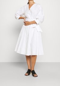 Robe enveloppante blanche avec manches bouffantes 3/4, tissu texturé et taille cintrée. Associée à des sandales noires à bout ouvert sur un fond gris.