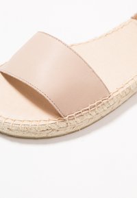 Beige lädersandal med bred rem, naturlig jutesula och sömdetaljer. Platt design, öppen tå och slät yta.